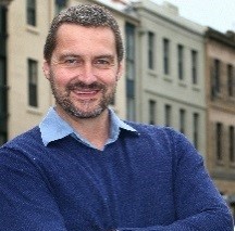 Image of a man in a blue jumper with old buildings behind him smiling at the camera.