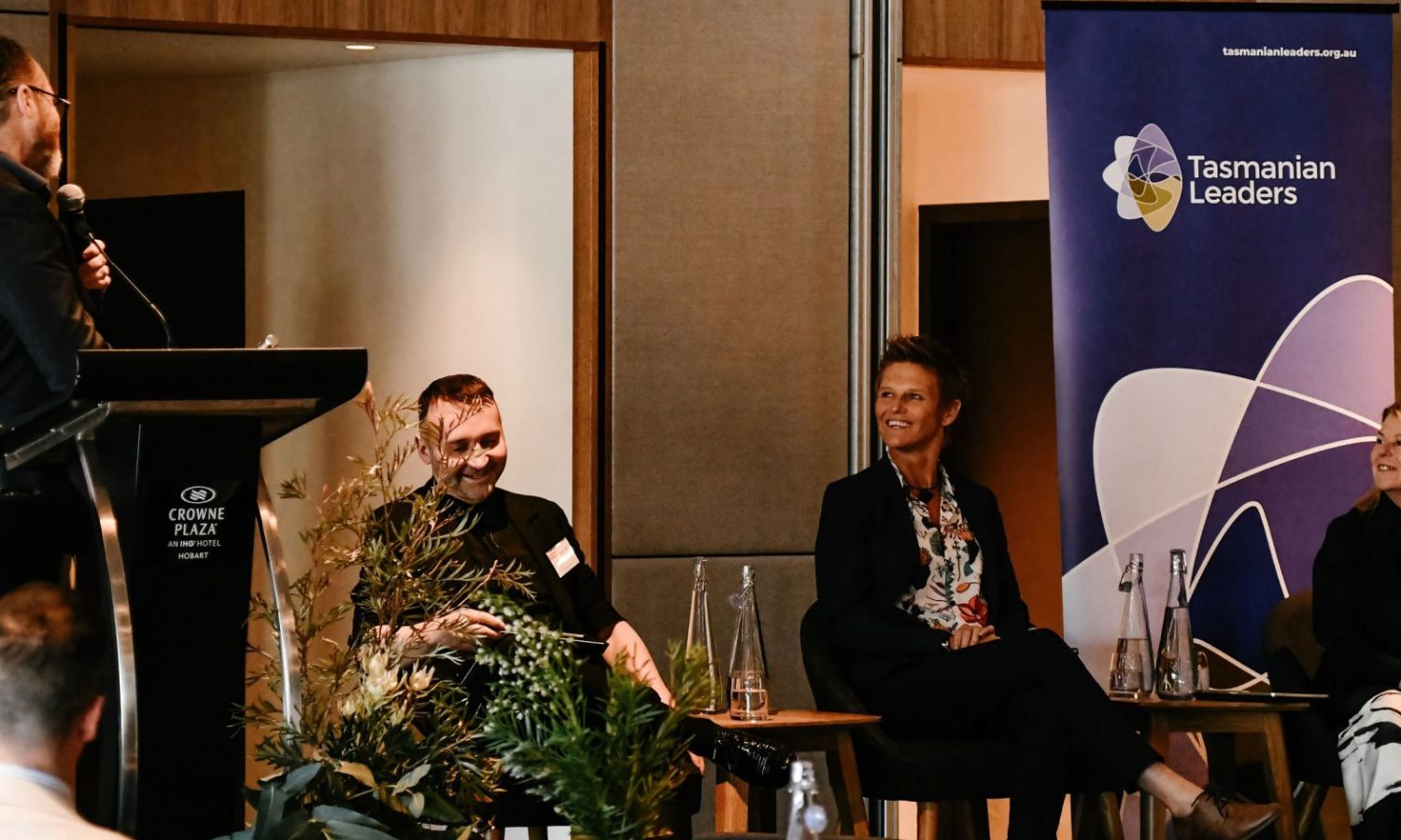 Photo of three panellists at the Symposium seated on a stage. They are all smiling and listening to facilitator.