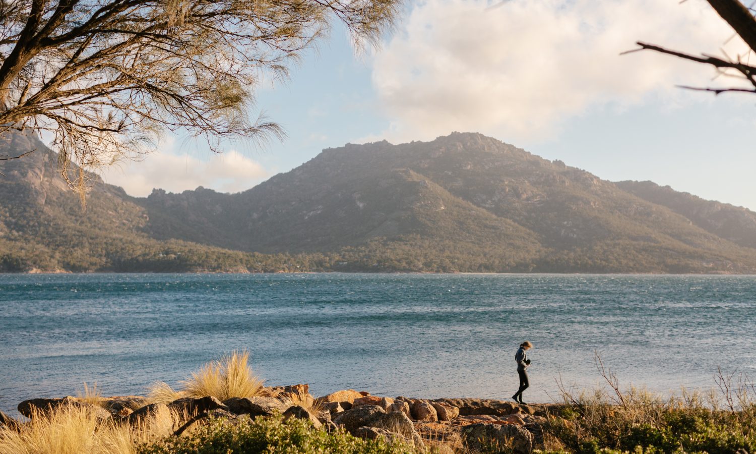 Image of a woman walking on rocks by the ocean with mountains as a backdrop.