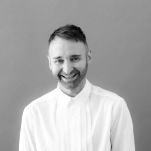 Image of a man in a white shirt standing against a grey background.