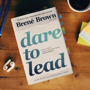 A book on a wooden table, surrounded by ear pods, a cup , post it notes and pencils. The book has coloured tabs on different pages.