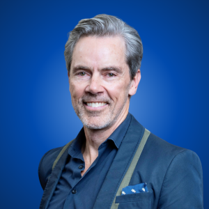 Portrait shot of Martin Beeche wearing a blue shirt and suit jacket with neat short hair, smiling at the camera with a blue background.