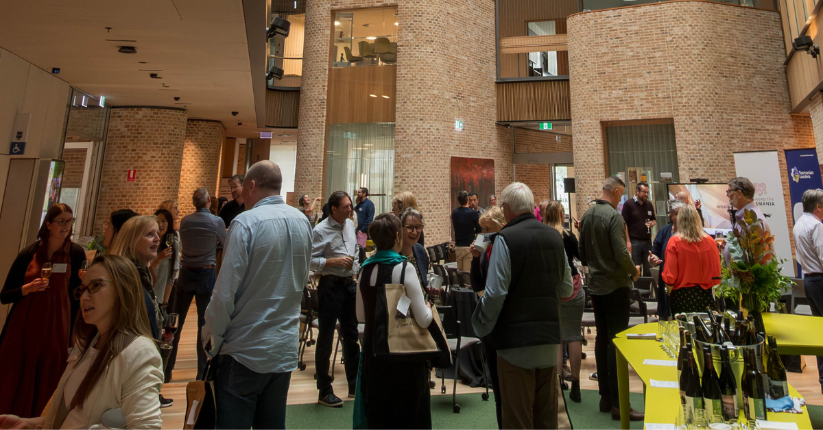 Image of participants of the Leadership Summit networking in the Rivers Edge Building at UTAS. they are all talking amongst themselves many are smiling . Tall internal brick walls are the back drop.