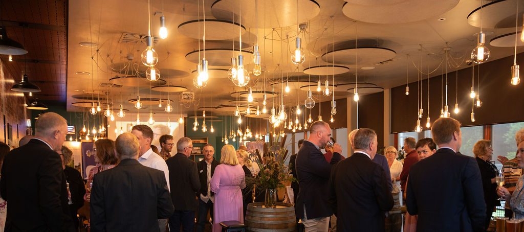 Landscape photo taken at the TLP18 Graduation. Lots of well dressed attendees networking with their backs to the camera, lots of warm light bulbs hang from the ceiling. There are wine barrels with foliage in the photo.