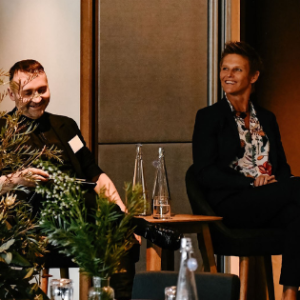 Panel photo of three panellists a man with short dark hair and a dark jumper, a woman with short spikey hair and a floral shirt and blonde woman wearing black and white. All are seated on a stage and smiling.