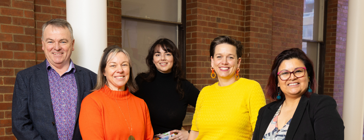 Landscape photo is of 5 2024 Tasmanian Leaders Participants standing indoors with a brick wall as backdrop. A man in a bright shirt and grey suit jacket, a woman in a bright orange roll necked jumper, a woman with long dark hair and a black jumper, a woman with a bright yellow jumper and bright orange earrings and a woman in a suit jacket, sark hair and pink glasses.