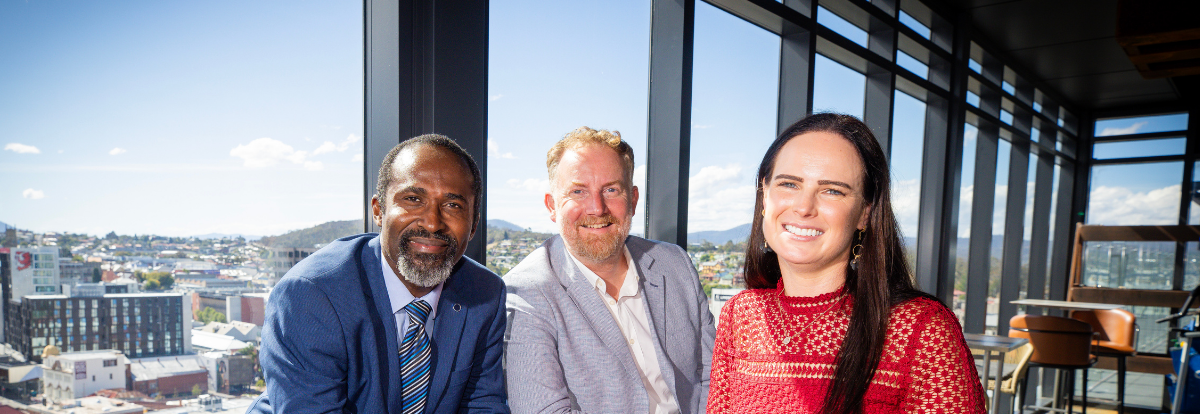 Head and shoulders shot of 3 TLP participants from 2023. A woman with long black hair and a long sleeved red dress, a fair haired man with a beard in a grey suit jacket and a dark haired man in a blue suit together. View behind them overlooks the city of Hobart.