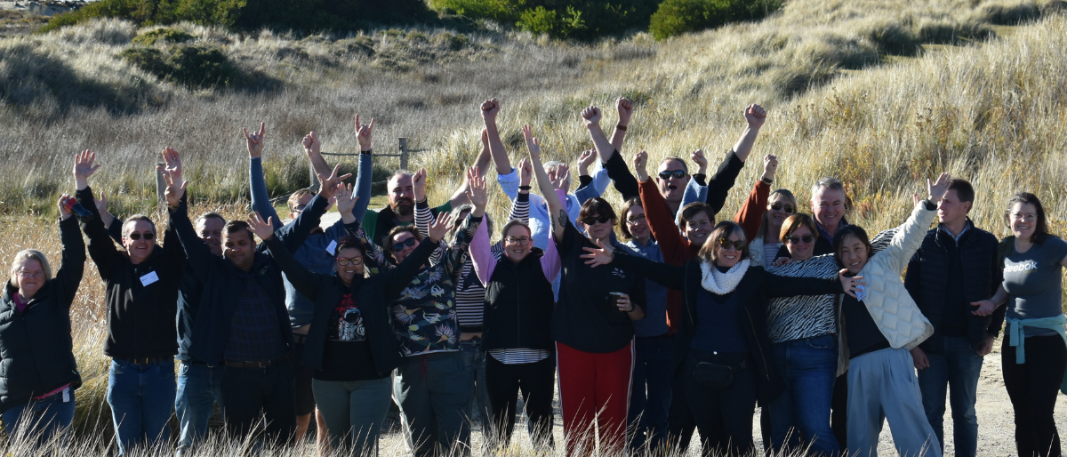 A landscape group photo of the 2024 Tasmanian Leaders Participants. Taken in a sand dune beach setting all in casual clothes with arms up in the air smiling at the camera.