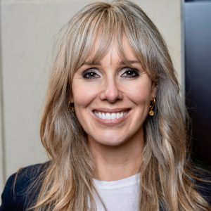 A close up head shot of Zoe Coyle facilitator of Dare to Lead. She has long blonde hair, smiling wearing gold hoop earrings, white t-shirt and blazer,