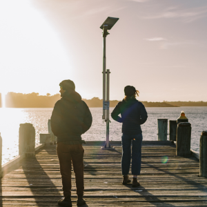 A cropped photo from Brand Tasmania showing two people walking towards the end of a pier. The first person is wearing a dark hoodie, brown cord trousers and Blundstone workboots and a beanie. The second person is wearing a hooded jacket, dark beanie and jeans with a cuff turned up and brown desert boots. Both people have their back to the camera and are looking in opposite directions. The sun is shining on the water on the left, and there is a slight wind. In the distance is a coastline with hills