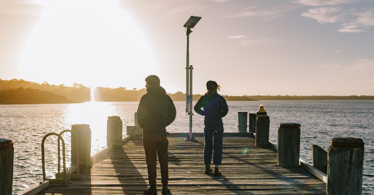 A cropped photo from Brand Tasmania showing two people walking towards the end of a pier. The first person is wearing a dark hoodie, brown cord trousers and Blundstone workboots and a beanie. The second person is wearing a hooded jacket, dark beanie and jeans with a cuff turned up and brown desert boots. Both people have their back to the camera and are looking in opposite directions. The sun is shining on the water on the left, and there is a slight wind. In the distance is a coastline with hills