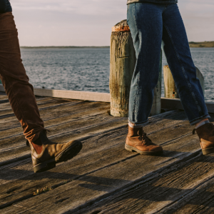 A cropped photo from Brand Tasmania showing two pairs of legs walking on a pier. The first pair of legs are wearing brown cord trousers and Blundstone workboots. The secong pair of legs are wearing jeans with a cuff turned up and brown desert boots.