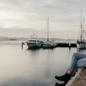 a woman is sitting on the edge of a wharf, looking away from the camera. In the distance there is a couple of boats moored at the wharf, and a mountain scape in the far background