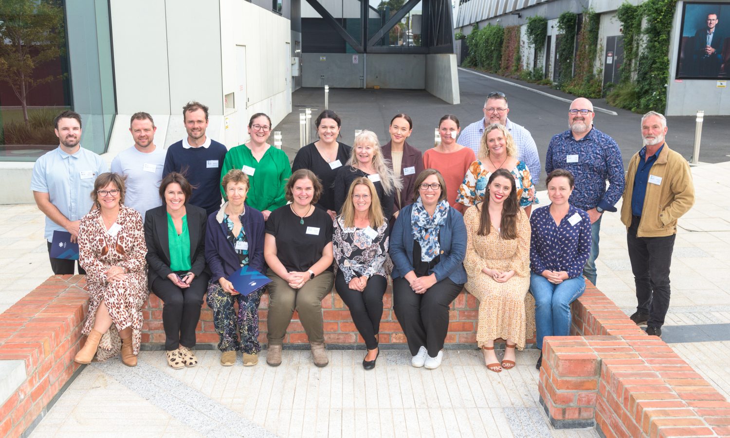 Group photo of the LARC program participants at their graduation event. A group of people are seated on a brick wall / standing behind a brick wall in front of a large educational campus building.