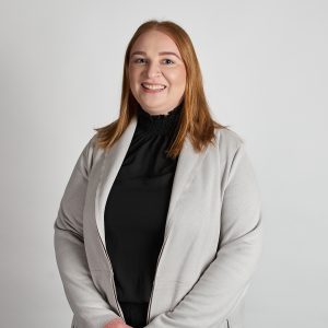 Portrait photo of El Chettle, graduate of I-LEAD Women in Industry 2025. El has mid length straight red hair, blue eyes and pale skin. She is standing, with hands clasped to the front. El is wearing a light grey jacket and high necked black blouse.