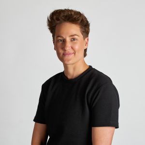 Headshot of Chey Sullivan, I-LEAD Women in Industry 2025 graduate. Chy has short thick red-brown hair styled high off her forehead, golden toned skin and blue eyes. She is wearing a plain black t-shirt and gently smiling at the camera.