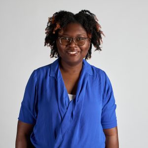 Portrait photo / headshot of Teresa Lemaire, graduate of I-LEAD Women in Industry 2025. Teresa has tight brown curly mid length hair with some orangey red tips, dark warm chocolate skin and brown eyes. She is wearing black framed glasses and a blue silk cross-over shirt, over a white camisole top. She is looking towards the camera straight on and smiling