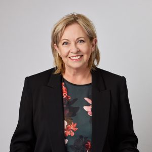 Portrait photo / headshot of Jillian Aylett-Brown. Jillian has mid blonde hair cut in a long tousled bob, blue eyes and pale skin. She is wearing a black suit jacket over a printed dark green top with large coral and pink floral shapes. She is smiling and looking towards the camera
