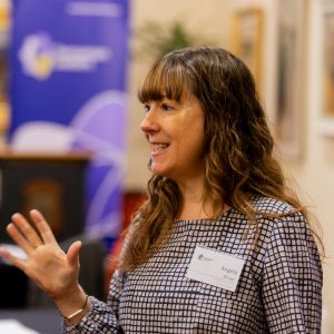 Image of Tasmanian Leaders Chief Executive Officer Angela Driver. Angela is looking to the left of the image with one hand raised for emphasis as she speaks. She has long wavy dark hair with blonde highlights, and is wearing a blue and white small check printed blouse and name tag.