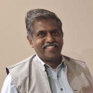 Headshot of Ajit Saralabai Ramadas, 2026 TLP Participant. Ajit has short greying dark hair worn parted to one side. He has dark brown eyes and brown skin, and wears a short salt and pepper moustache. He is smiling with teeth at the camera, wearing a pale blue business shirt and herringbone patterned Nehru waistcoat