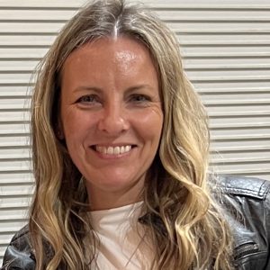 Portrait photo of Amanda Hardinge, 2026 TLP Participant. Amanda has dark blonde greying wavy long hair, worn in a centre side part, with hazel eyes and slightly tanned skin, She is wearing a weathered leather jacket over a white top and smiling with teeth towards the camera