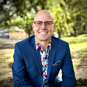 Portrait photo of Benny Callaghan. Benny is wearing a rroyal blue jacket, retro floral patterned shirt and glasses, with a bald head and a big smile. Benny is sitting in a park with dappled light coming through the trees behind him