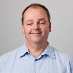 Headshot of David Griffiths, 2026 TLP Participant. David has short dark hair worn cropped close with a widow's peak, brown eyes and pale skin, wearing a pale blue business shirt worn with the top button open. He is smiling at the camera.