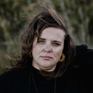 Portrait of Emily-Rose Wills, 2026 TLP participant. ER is standing in a windy landscape, with her fine dark brown shoulder length hair being blown away from her forehead. She has grey eyes and pale skin with light freckles. ER is wearing a black long sleeved loose top, large brown marbled round hoop earrings, and a black cord necklace. She has one hand bent behind her head as if she has just brushed her hair aside