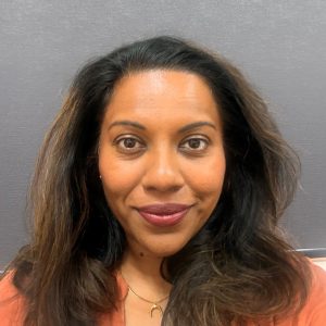 Headshot portrait of Ganga Fraser, 2026 TLP Participant. Ganga has long highlighted thick brown wavy hair, worn brushed away and to the side of her face. She has dark brown eyes, honey skin and large lips. Ganga is wearing an orange blouse and gold chain necklace with pendant