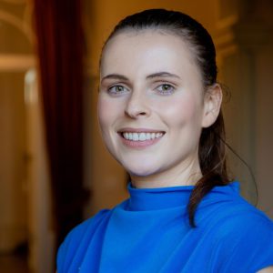 Headshot of Gemma Smith, 2026 TLP participant. Gemma has fine dark hair pulled back from her forehead to the back, light brown eyes and pale skin. She is gently smiling showing teeth and is gazing towards the camera. Gemma is wearing a cropped sleeve azure blue top with stand up neckline.