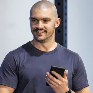 Headshot photo of Hamish McInerney, 2026 TLP Participant. Hamish has a shaved head, dark brown eyes, golden skin and slight stubble with a dark short moustache. He is wearing a blue t-shirt with a corporate logo slightly obscured. In one hand he is holding a smartphone, and is gesturing with an open hand towards the viewer.