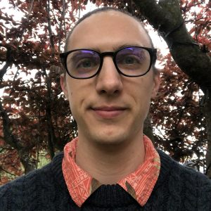 Portrait of Malcolm Johnson, 2026 TLP Participant. Malcolm has dark hair worn pulled back into a low ponytail, dark blue grey (?) eyes and honey pale skin. He wears black round framed glasses, a blue cable knit jumper over a red and orange printed shirt collar. He is standing in front of a large tree with red leaves that is filtering the light behind him