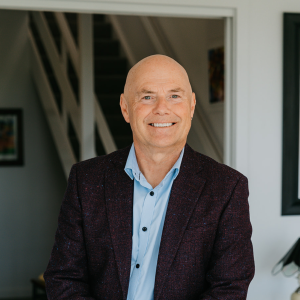 Portrait photo of Michael Johnstone, 2026 TLP participant. Michael has a bald head, blue eyes and slightly tanned pale skin. He is wearing a deep brown / blue tweed suit jacket over a pale blue business shirt with an open collar.