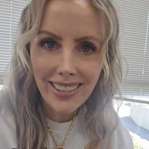 Close up headshot of Samantha Sullivan, 2026 TLP Participant. Samantha has long wavy silvery blonde hair, half worn up in a messy bun, with long wavy fringe framing her face. She has blue eyes and pale skin. Samantha is wearing a white windcheater with a print of two people on the front, with a chunky gold necklace with heart pendant over the top. She is leaning towards the camera and smiling with teeth.