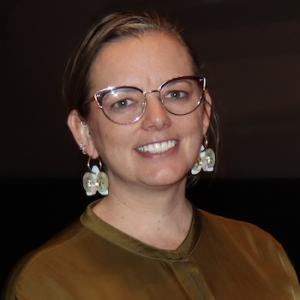Headshot photo of Siobhan Mackay, 2026 TLP Participant. Siobhan has dark blonde hair worn up and brushed to the side, green / brown (?) eyes and mid toned skin. She is wearing black and bronze metal cats eye framed glasses, teal and mother of pearl on hoop earrings, and a bronze silk blouse.