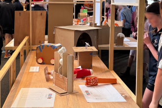 Photo of part of the NARLE project installation in George Town gallery. A long timber table with a frame around two sides is in the middle ot the image. On top of the table are 6 prototype models made of cardboard of various shapes. at the back of the table is another open timber frame, with a table behind that holding more prototypes and panels. to the sides and back of the image is a blurred group of people including adults and school children