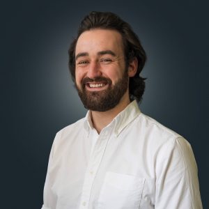 Portrait of Liam Kearns. Liam is a young man with wavy dark hair brushed back from a high forehead. He has a thick dark beard and moustache, and brown eyes. Liam is wearing a plain white business shirt with the collar unbuttoned.