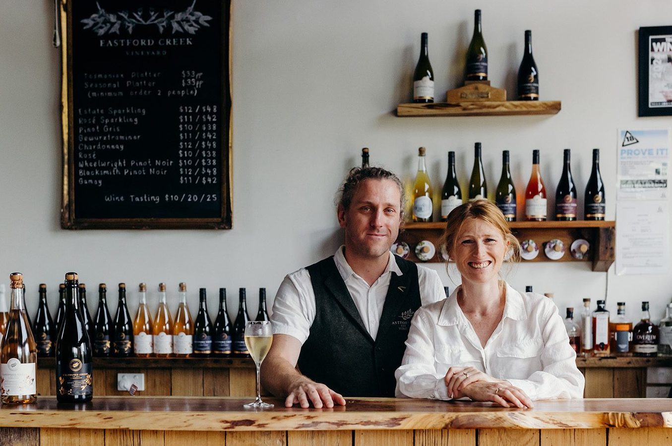 Two people smiling behind the timber cellar door counter at Eastford Creek Vineyard, surrounded by bottles of Tasmanian wine.
