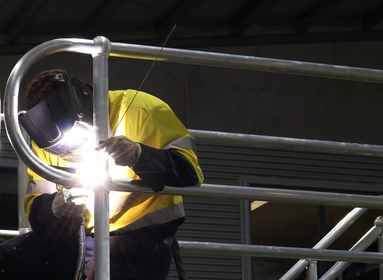 Worker in high-visibility clothing and protective welding equipment fabricating a large metal structure in an industrial workshop.