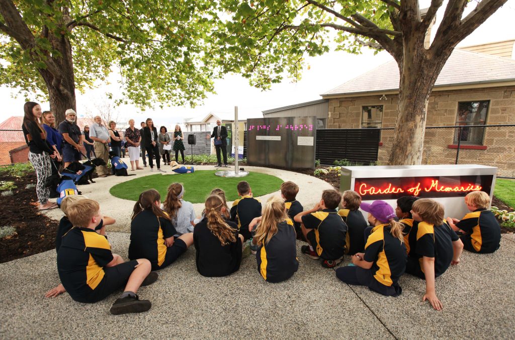 School children and community members gathered outdoors for the opening of the Garden of Memories, with illuminated artwork installed among the garden beds.