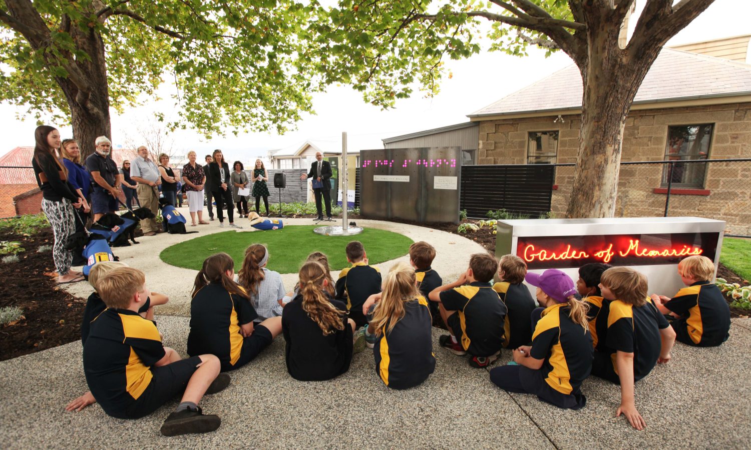School children and community members gathered outdoors for the opening of the Garden of Memories, with illuminated artwork installed among the garden beds.