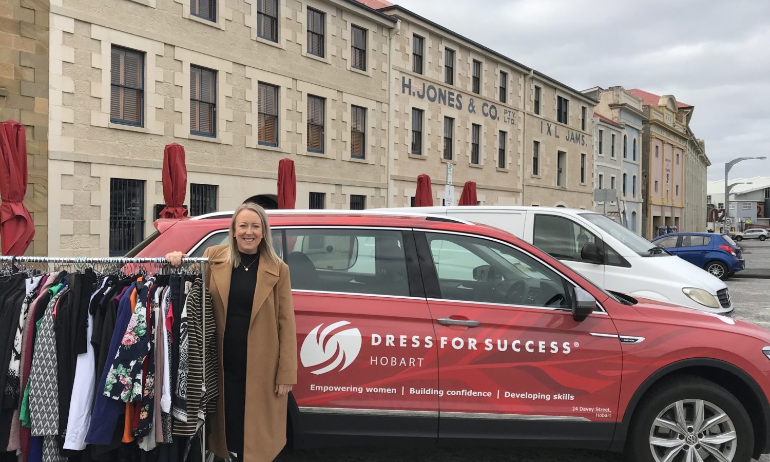 Woman standing beside a rack of clothing and a Dress for Success Hobart vehicle outside a heritage building.