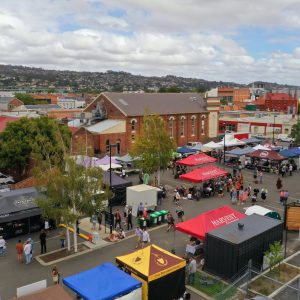 Elevated view across a busy Tasmanian community market, with people gathering among food stalls and colourful marquees surrounded by city buildings.
