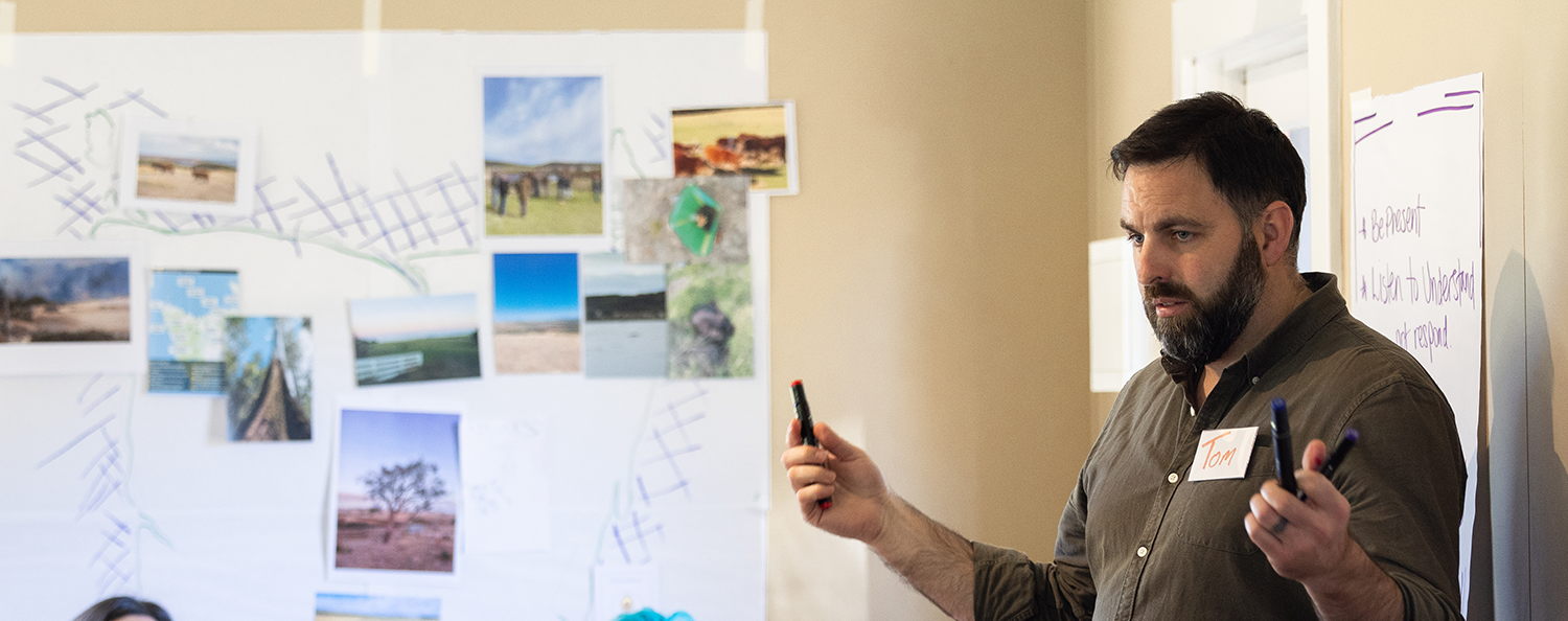 A facilitator speaking to a seated group during a Tasmanian Leaders retreat, with workshop notes, maps and photographs displayed around the room.