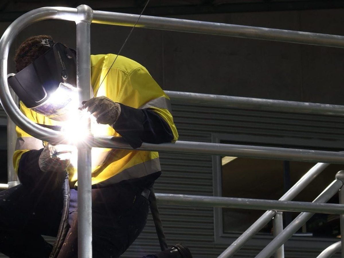 Worker in high-visibility clothing and protective welding helmet welding a stainless steel structure inside an industrial workshop.