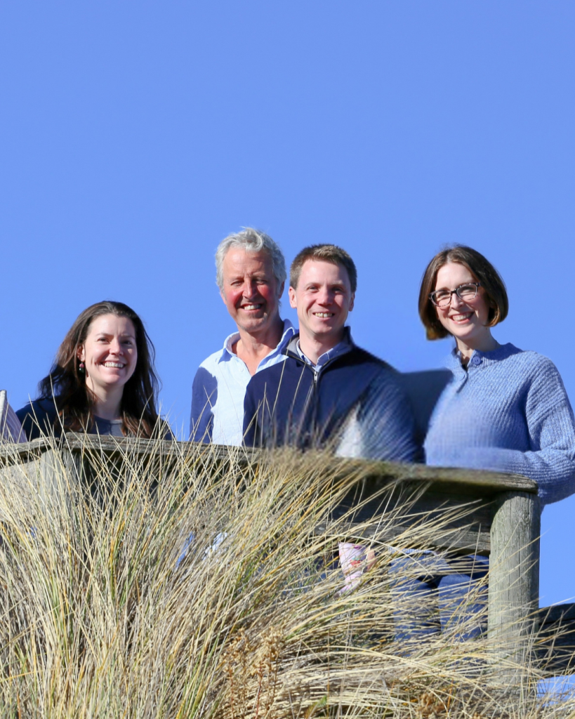 Four people smiling outdoors behind a timber fence, with coastal grasses in the foreground and a clear blue sky behind them.